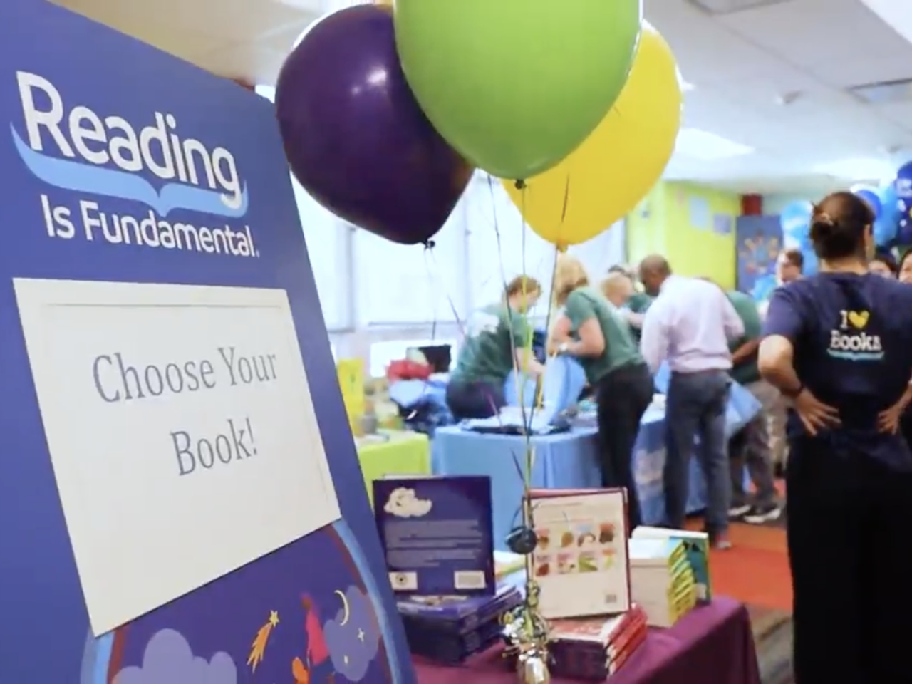 A sign that reads "choose your book!" on an event stand with balloons 