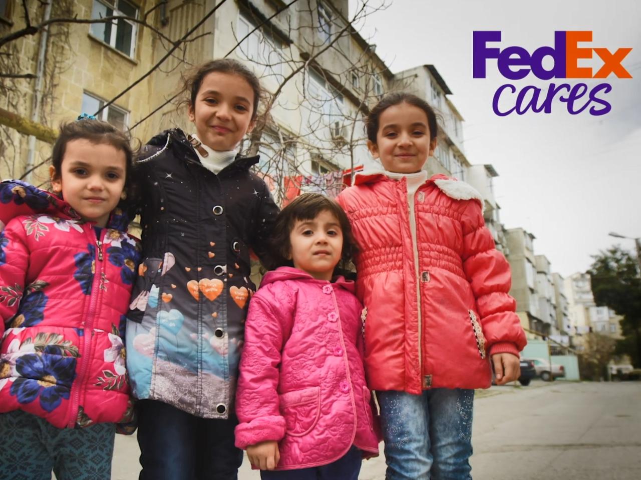 Four children posed close together, smiling outside residential buildings. "FedEx Cares" in the corner.