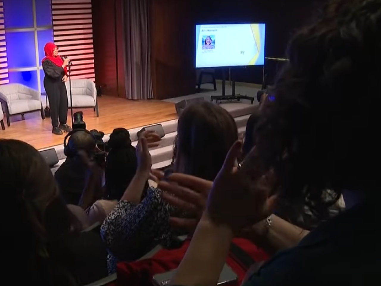 Saleemah Abdul-Ghafur speaking on a stage with three armchairs. An audience in front of her.
