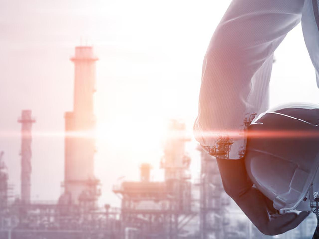 A person holding a hard hat, smoke stacks in the distance.