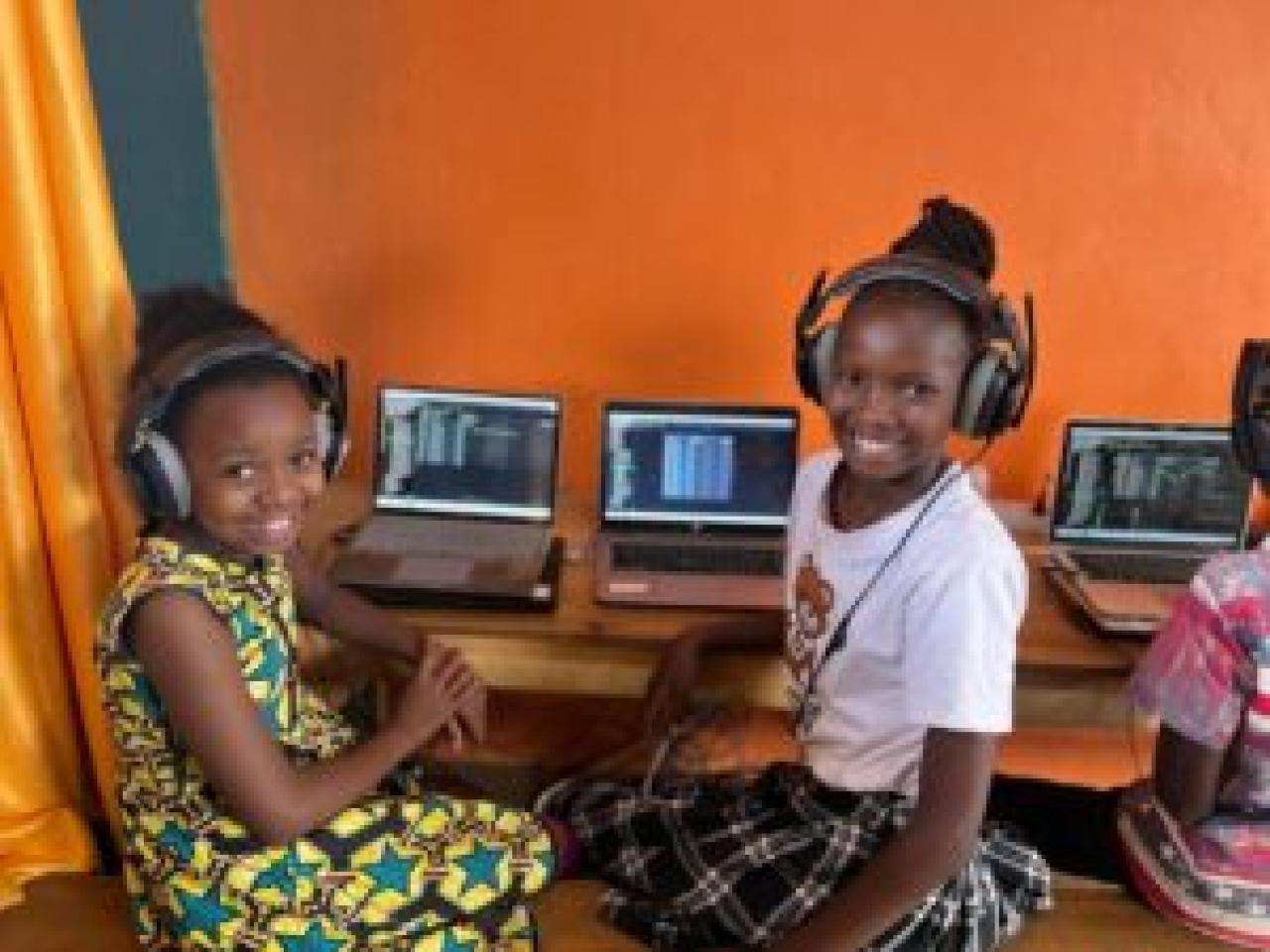 Two smiling children with headphones at a desk with open laptops.