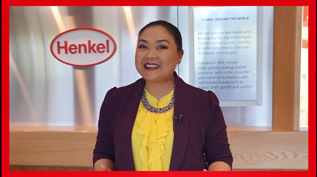  A person in front of a Henkel sign, smiling.