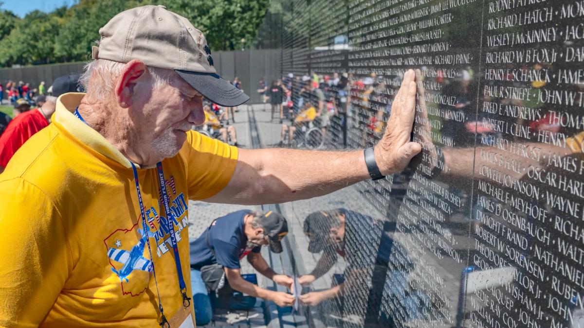 A person with a hand on a memorial wall covered in names. Their eyes are closed and head bowed.