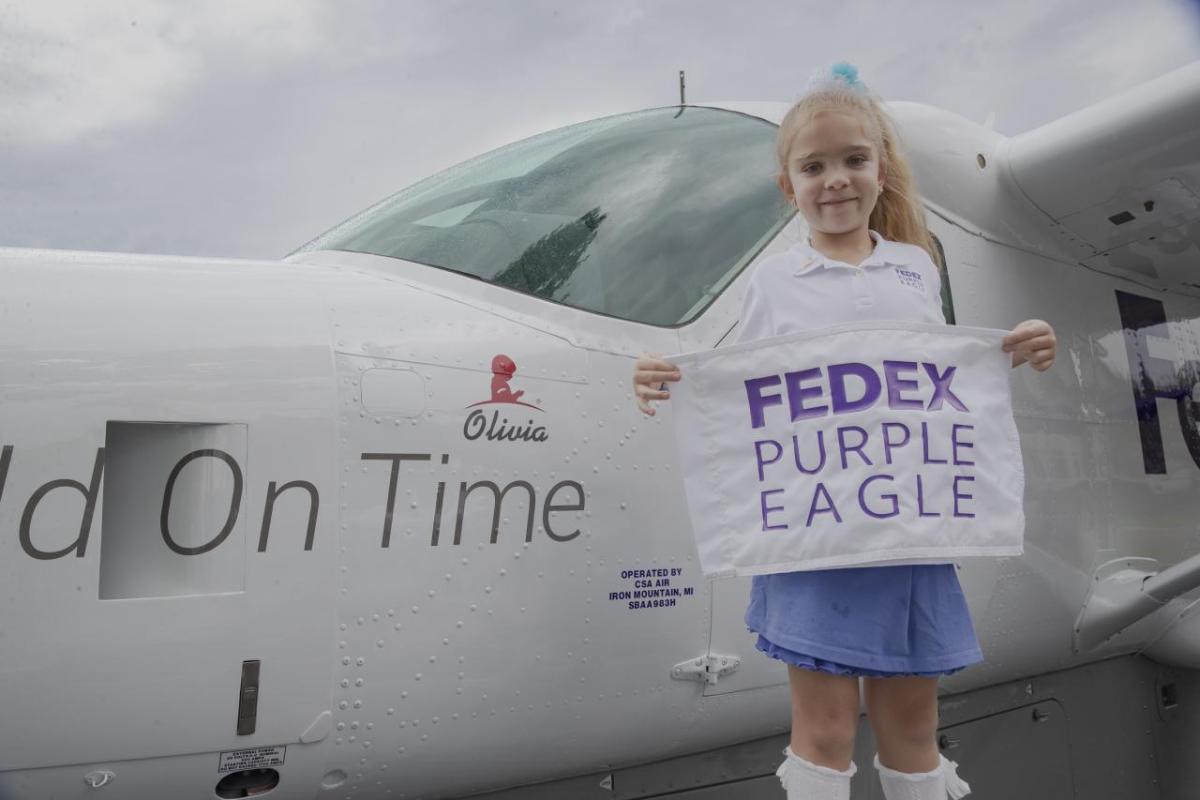 Olivia standing next to the plane with her name on it holding a "FedEx Purple Eagle" sign.