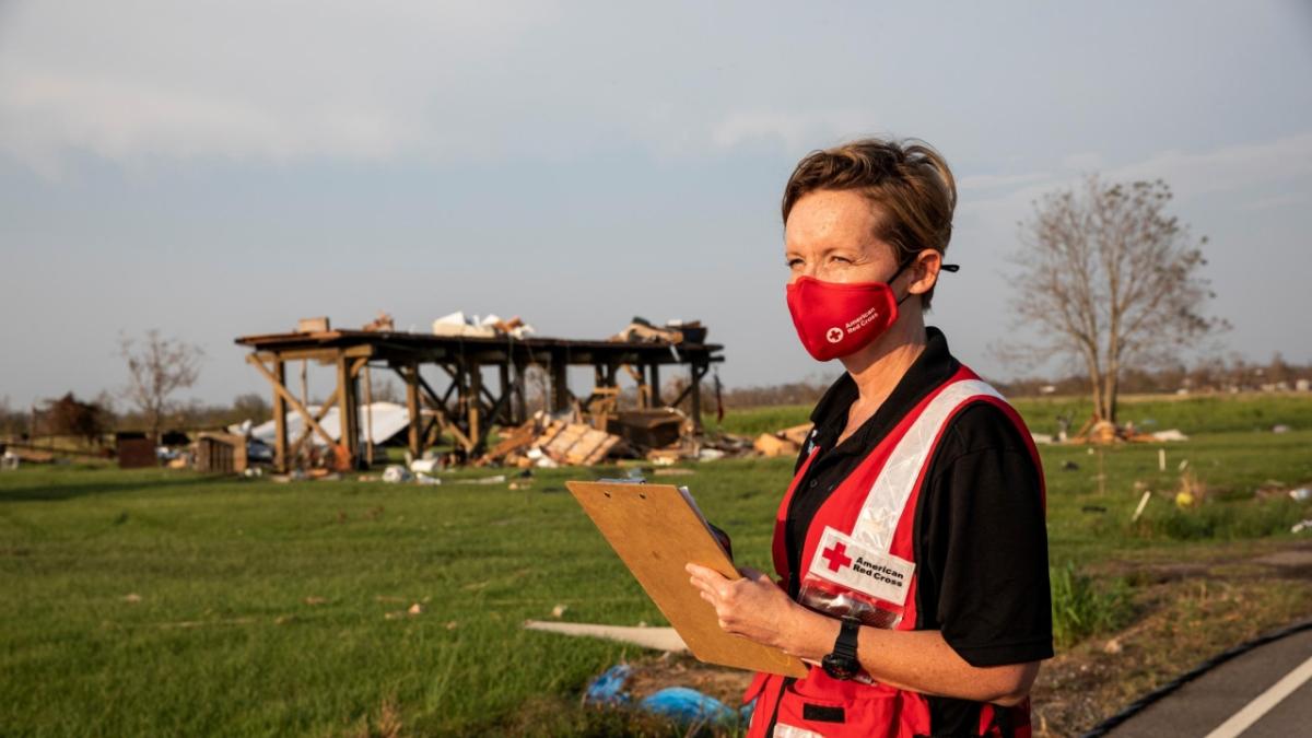 Red Cross volunteer carries a clip board in a field with debris scattered