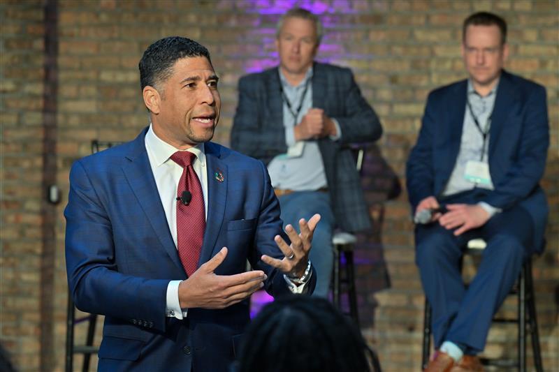 man speaking with two men behind him in chairs 