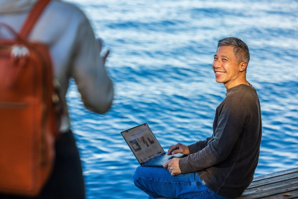 Person on a laptop seated next to a lake.