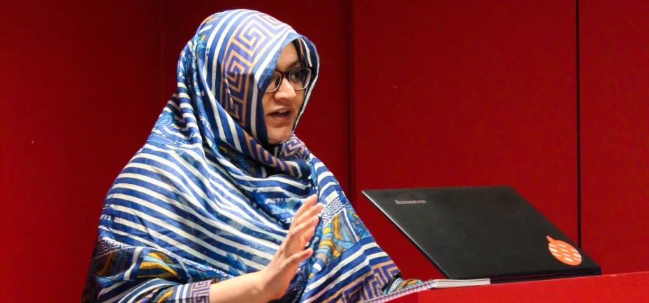 Faiza Yousuf standing at a podium with a laptop. Red walls behind her.