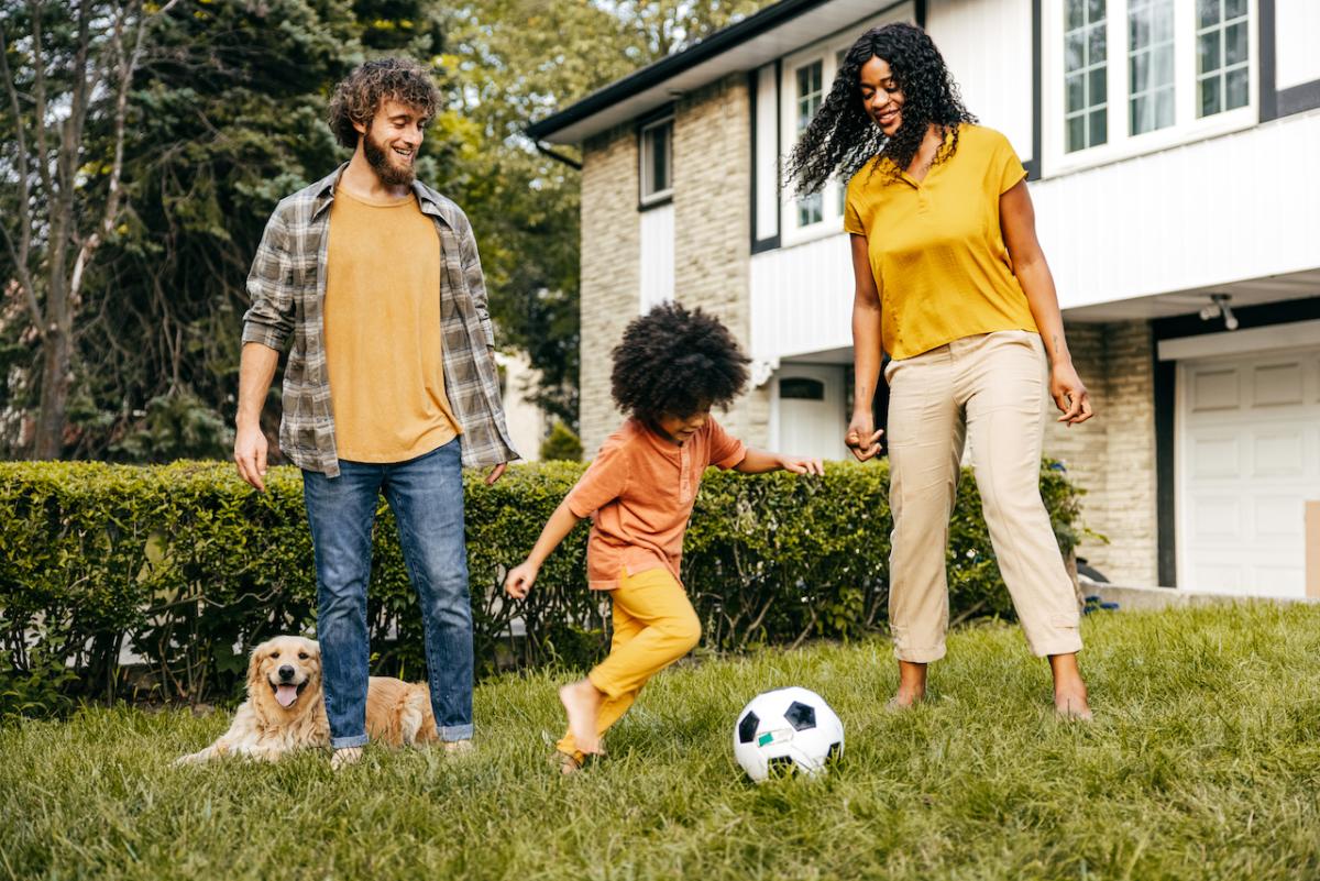 Parents playing soccer with child and dog outside of house