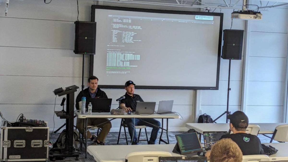 Two people seated at the front of a room, a display behind them with computer code on it.