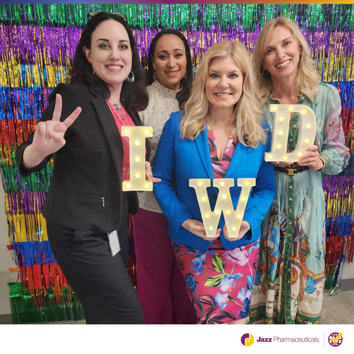 Four people posed in front of a wall of colorful fringe. Three holding "IWD" letters.