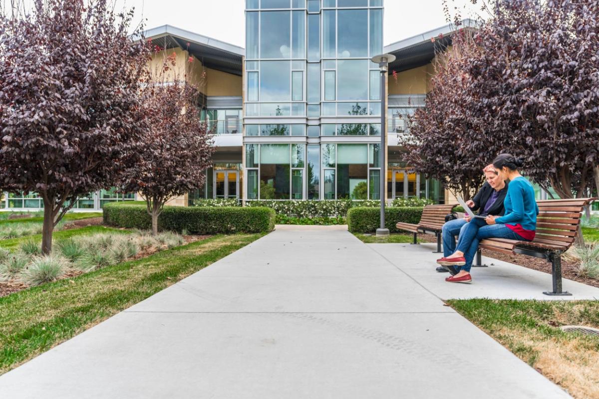 Two people sitting on a bench outside a building in a park like setting.