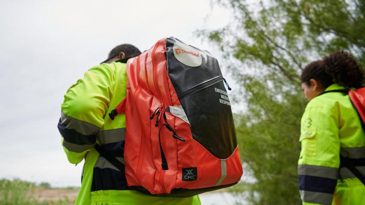 Two people wearing high-vis jackets and backpacks walking outside.