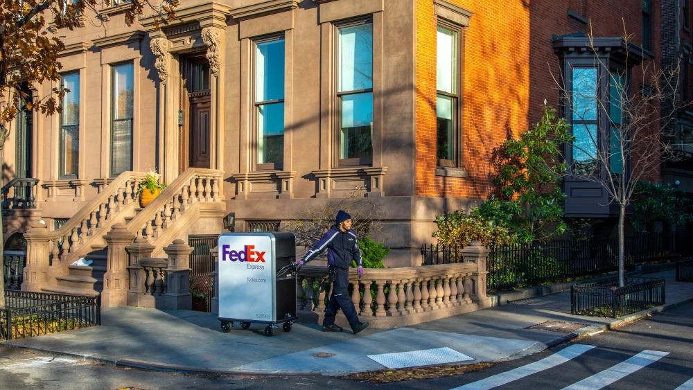 A person in FedEx uniform pulling a tall cart with FedEx Express logo on it going around a corner on the sidewalk in front of a house.