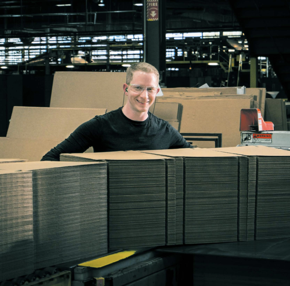 worker in a black shirt and safety glasses standing behind stacks of cardboard