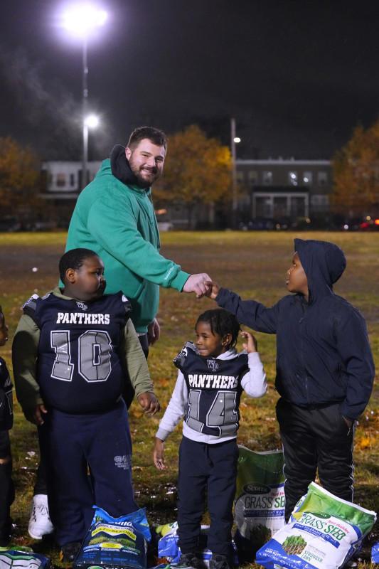 Landon shaking a childs hand on a football field, two others in uniform and bags of grass seed on the ground.