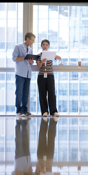 Two people standing in an open room with tall windows, looking at the same paper.