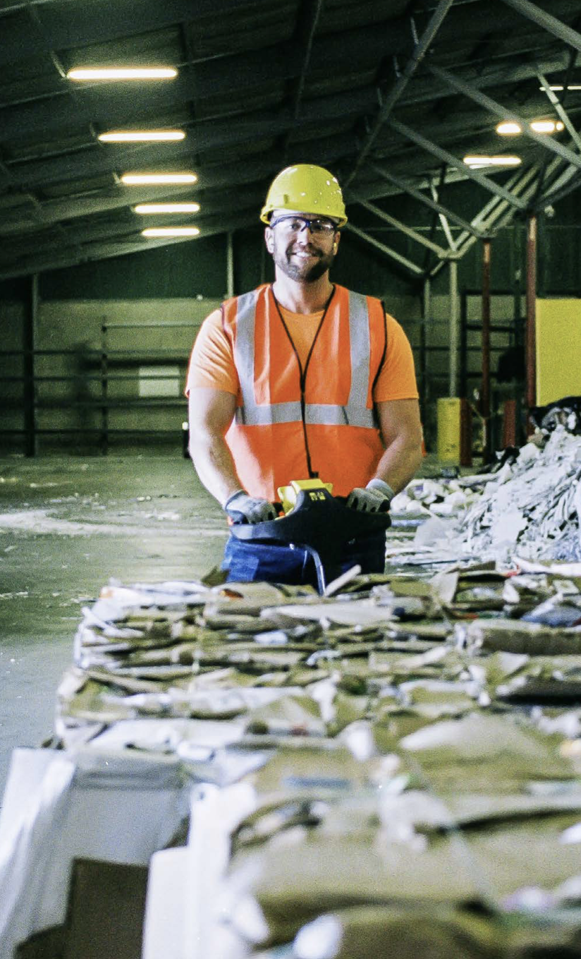 Worker in an orange safety vest and yellow helmet standing behind bales of paper recycling and smiling