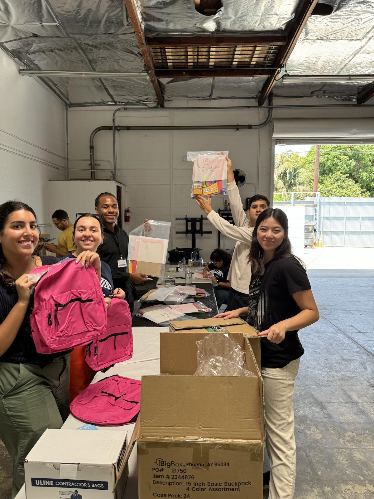 Volunteers standing around long tables holding boxes, pink backpacks, and other items for care packages