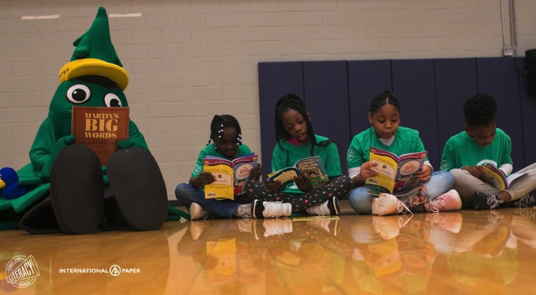 Children sitting in a line on a gym floor, a tree mascot next to them, all reading books.