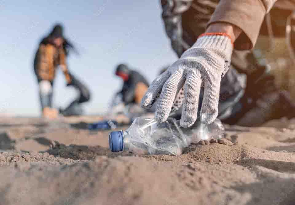 A gloved hand reaching down to pick up an empty plastic bottle on a beach. Others blurred in the background.