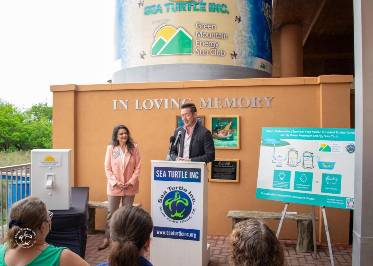 Man in casual suit addresses audience from podium, next to diagram poster, with large silo in the background