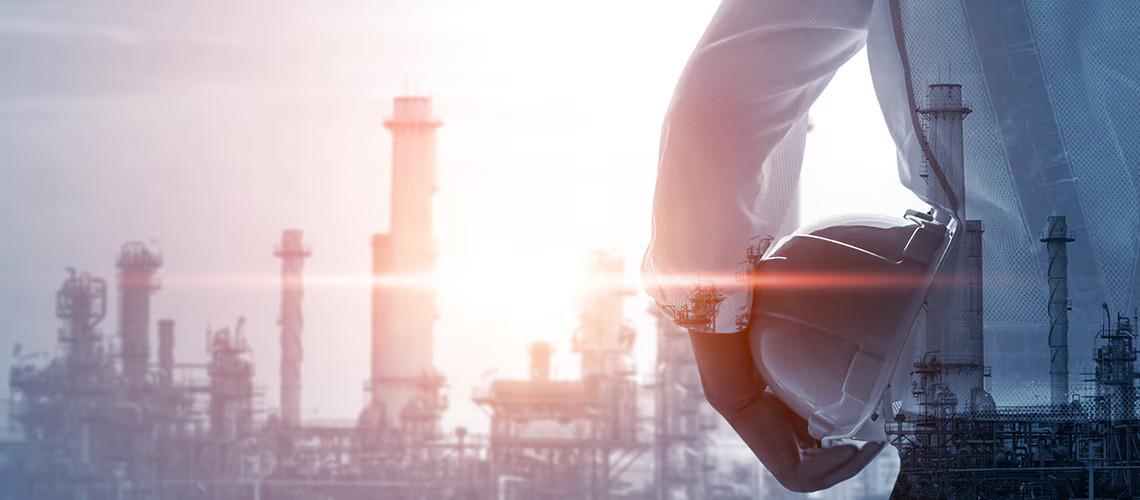 A person holding a hard hat, smoke stacks in the distance.