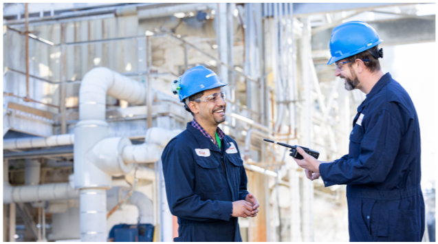 Two smiling people facing each other, wearing hard hats in an industrial area.