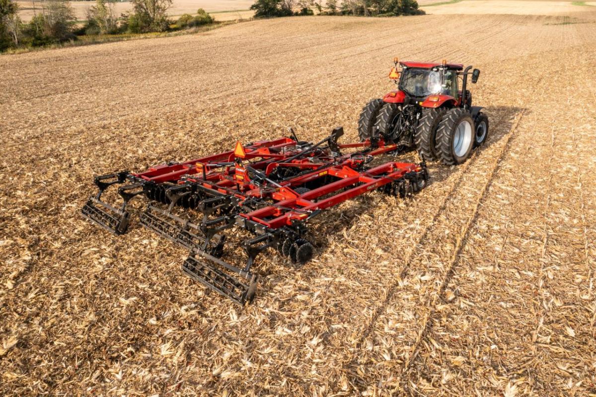 A large red tractor and attached tilling tool on a large harvested crop field.