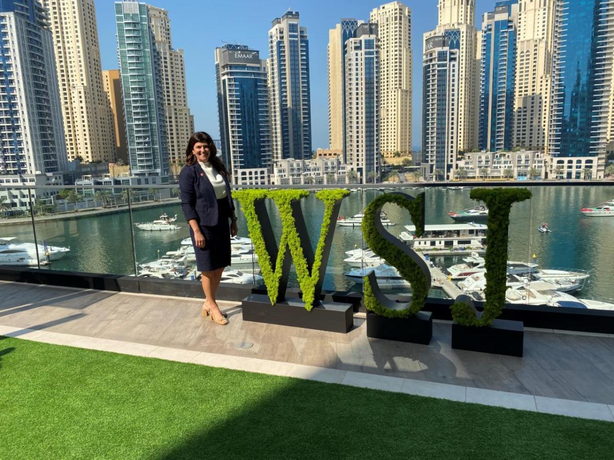 A person standing on a balcony next to green letters "WSJ" overlooking a body of water and skyscrapers.