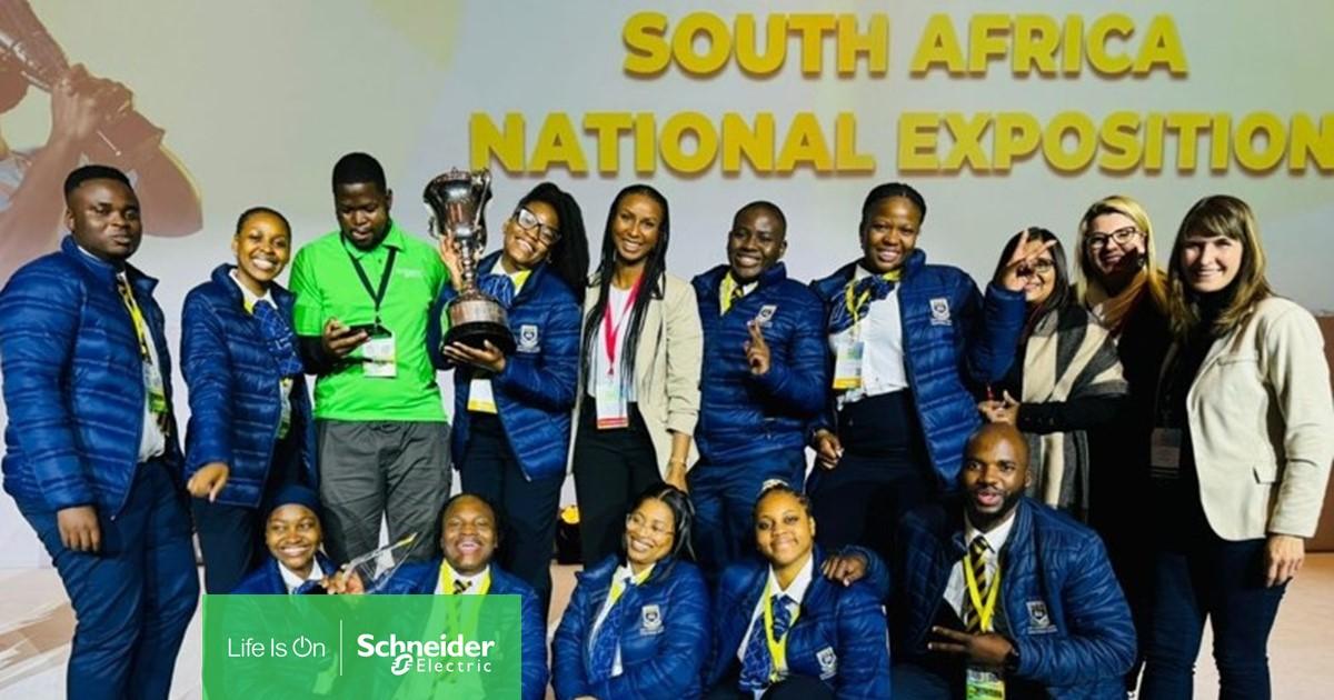 A group posed together, one holds a trophy and others a sign with Schneider Electric logo. "South Africa National Exposition sign behind them.