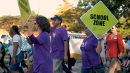 3 people in purple FedEx shirt, 2 of which are holding neon yellow "School Zone" signs