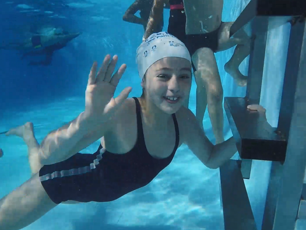 A child smiling and waving underwater in a pool, others swimming behind them.