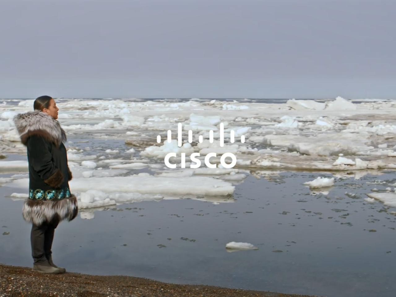 A person in warm clothing standing on a shore looking out over open water with chunks of ice and snow floating in it. "Cisco" overlaid.