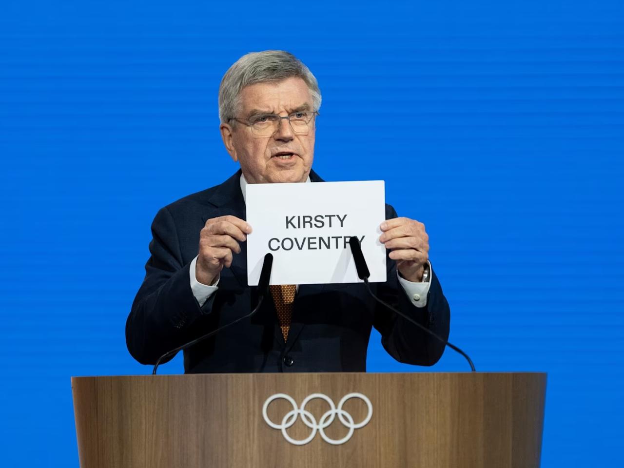 Man at lectern holding up s sign reading, "Kirsty Coventry"