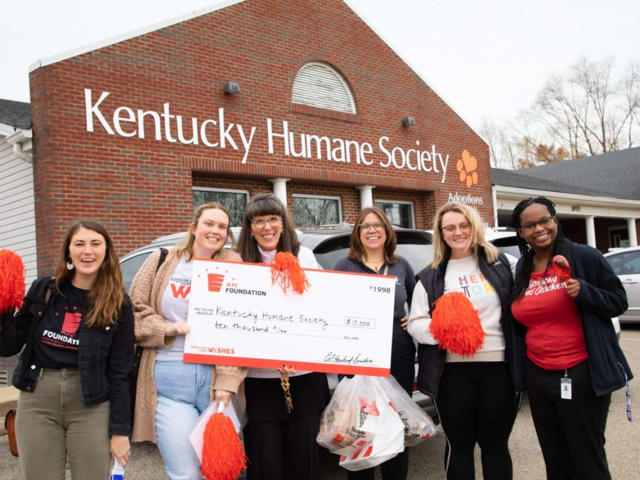 Six people standing together outside in front of the Kentucky Human Society, holding a large check