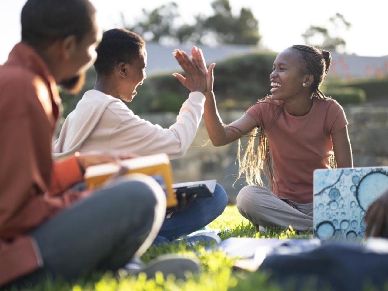 Photo of two students high fiving while sitting on the grass