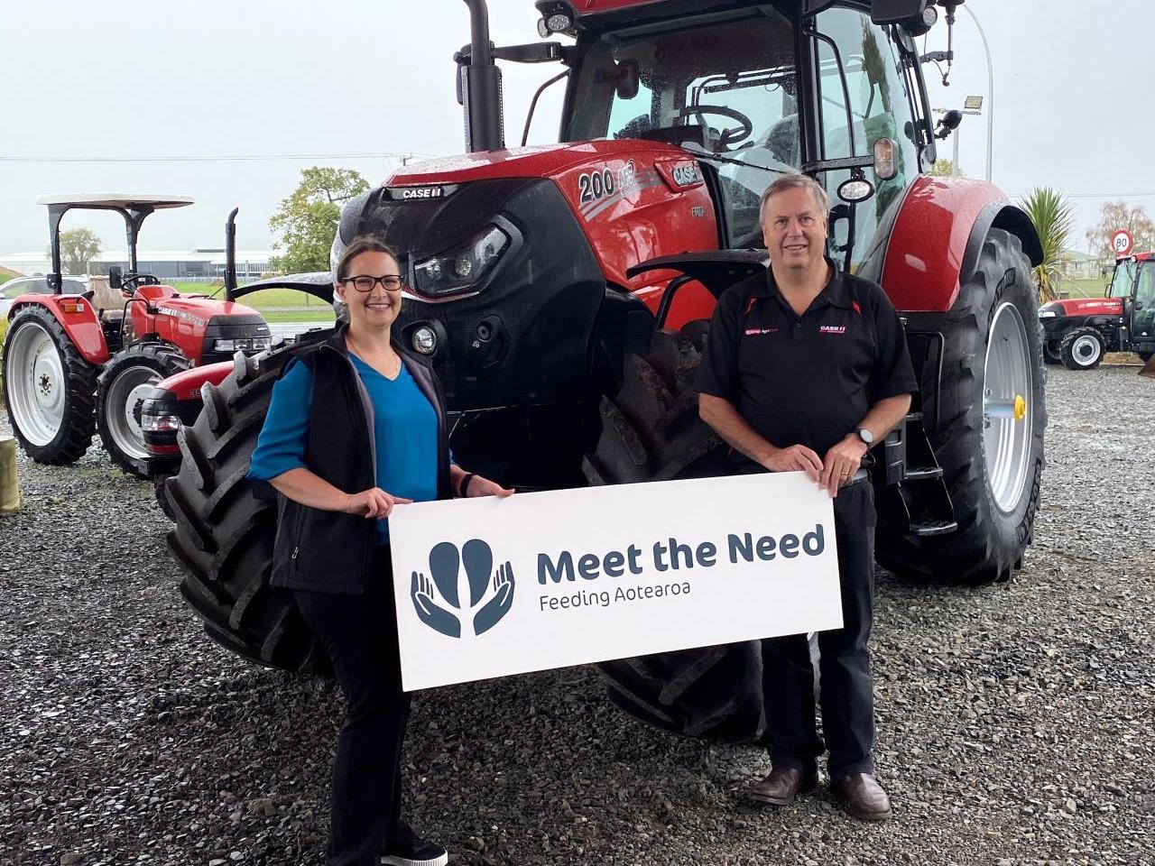 Two people stand in front of a tractor with a "Meet the Need" sign