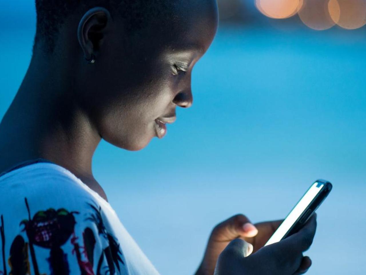 Close-up of a young woman using a smartphone at dusk, her face illuminated by the screen’s glow. She appears focused, with a soft blue background and blurred city lights in the distance.