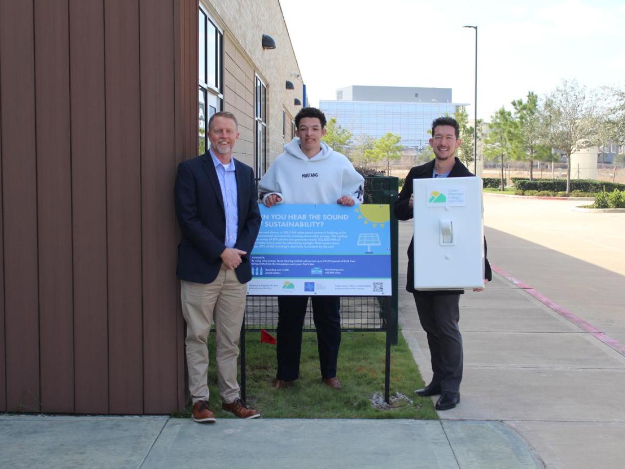 Three men standing around blue sign smiling, with one of them holding an oversized prop light switch