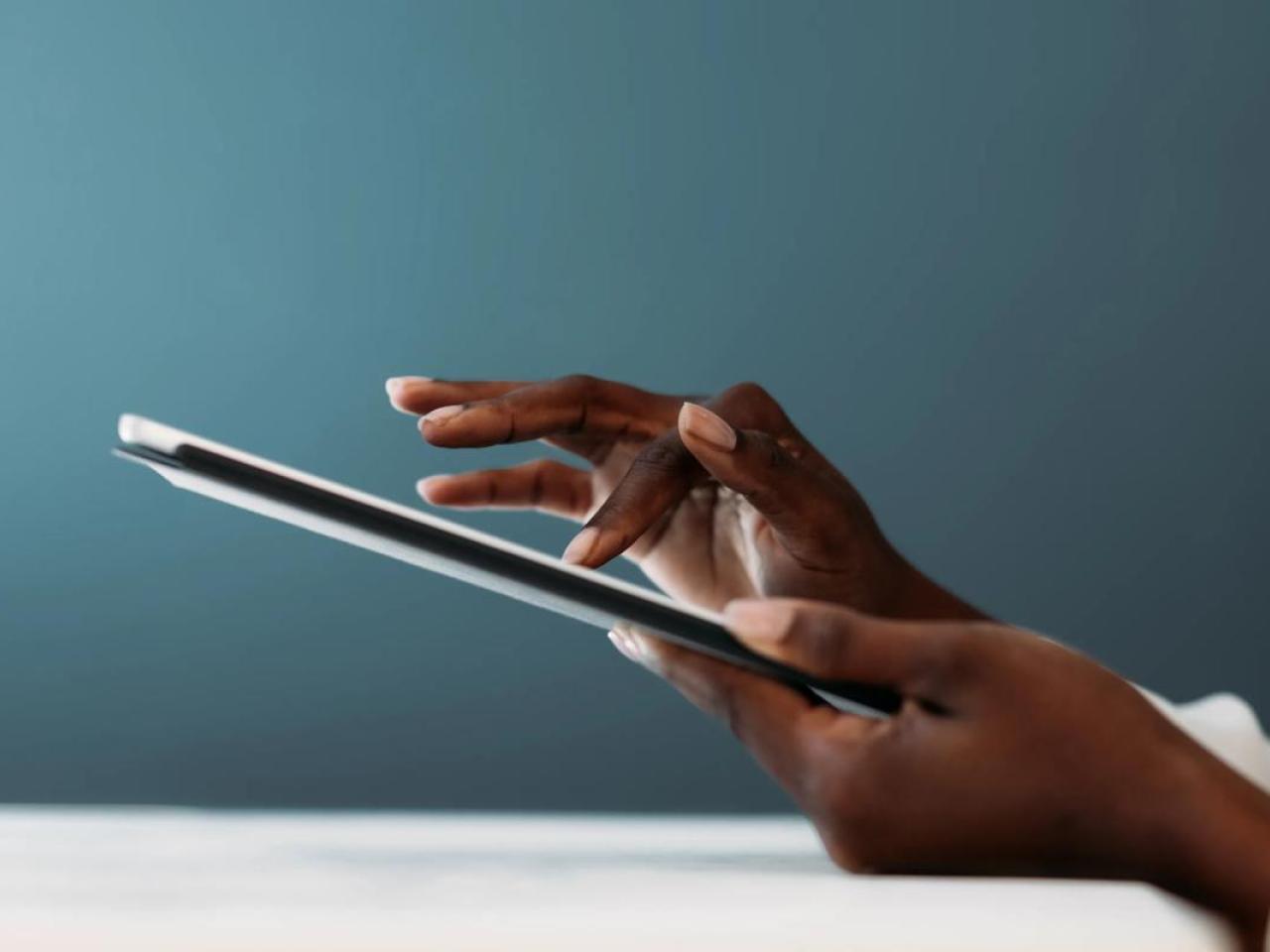 Close-up of a person’s hands using a digital tablet, with fingers gently tapping the screen against a soft blue background.