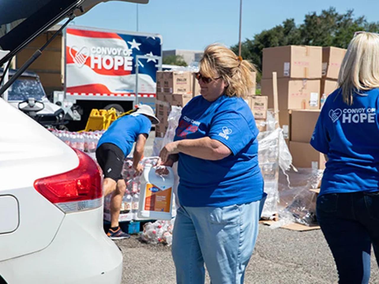 Volunteers loading the trunk of a vehicle. "Convoy of Hope" sign behind them and stacks of boxes.