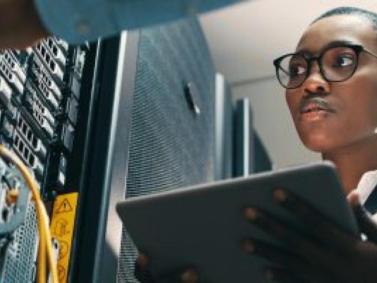 A person holding a tablet, looking at towers of electronic equipment.