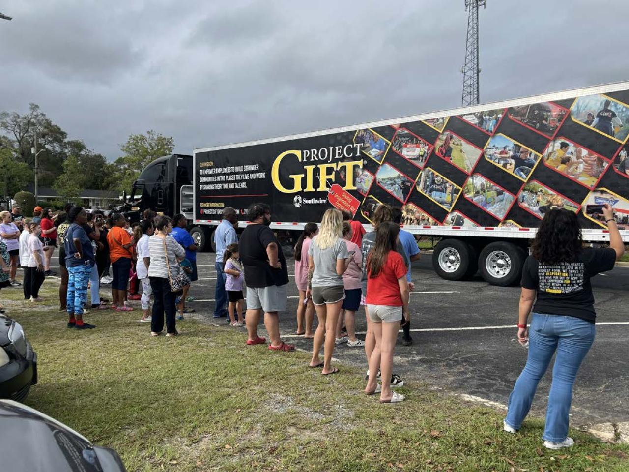 People waiting on a grassy area, looking at a semitruck with "Project Gift" on the side.