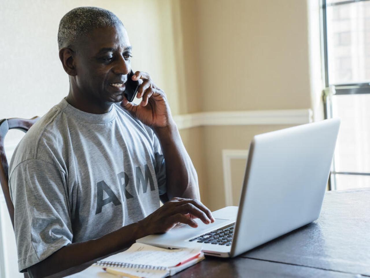 A person in an Army t-shirt on the phone while typing on a laptop