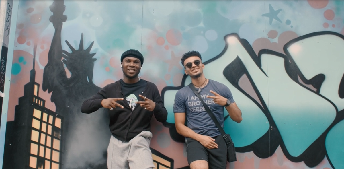 Two people leaned on a wall, both give a 'peace' sign. THe wall has a mural of the statue of liberty and other writing.