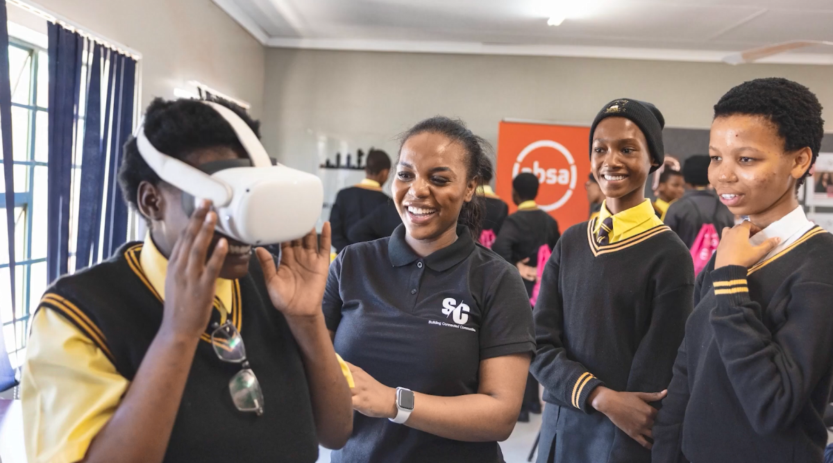 An adult assists school children using a VR headset.