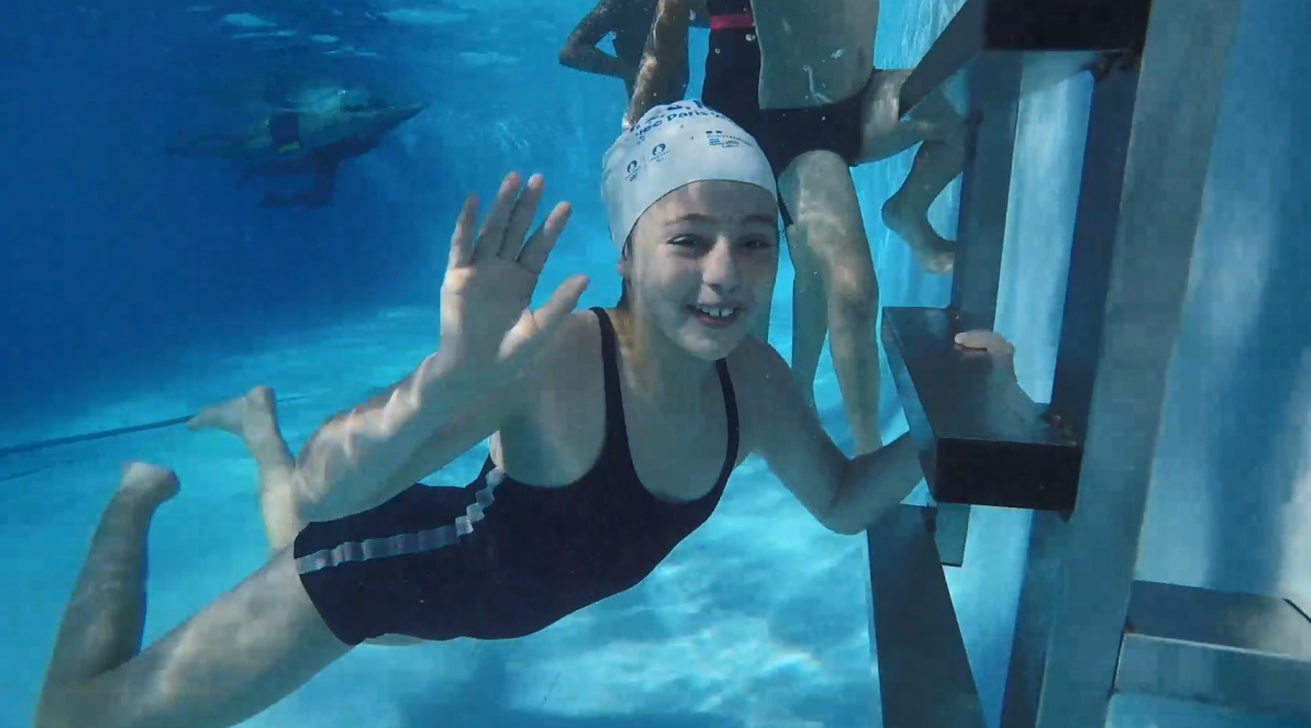 A child smiling and waving underwater in a pool, others swimming behind them.