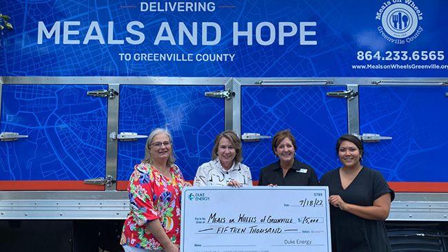 Four people in front of a blue background hold a large check to Meals on Wheels