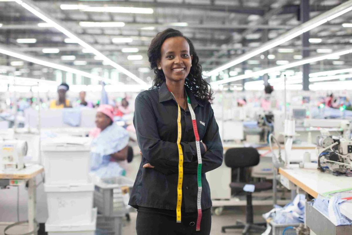 A young women in black clothing stands in front of a large room of women working on sewing machines. 